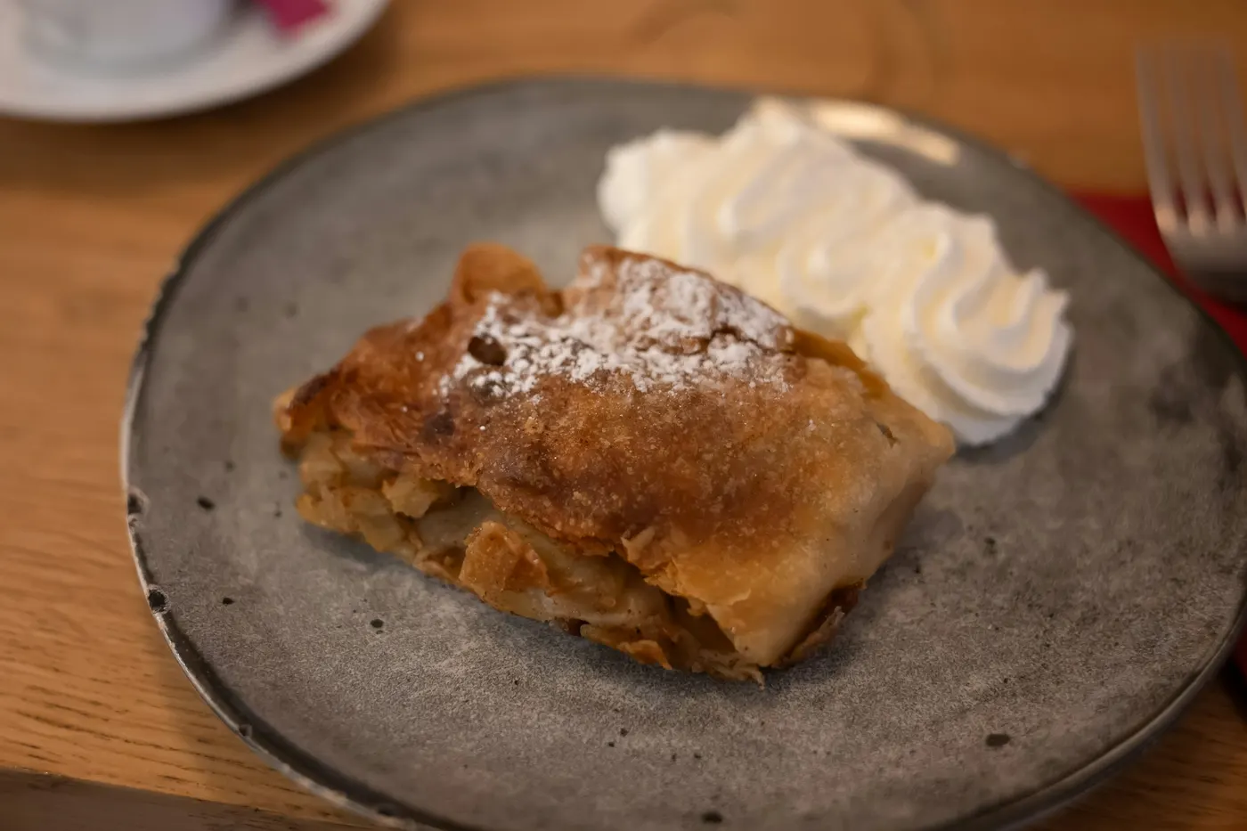 Ein Stück Apfelstrudel mit Staubzucker und Schlagobers auf einem grauen Teller